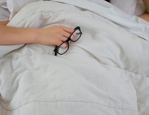 Woman in bed holding reading glasses with pillow covering part of her face